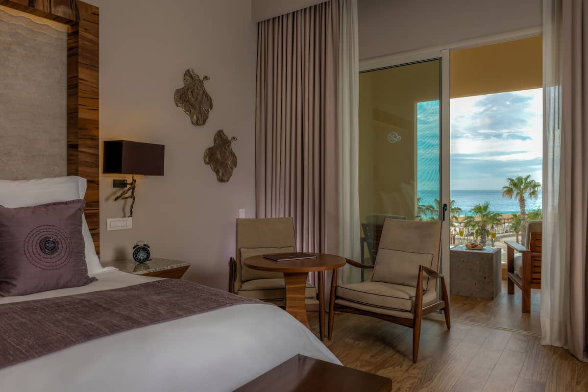 A hotel room with a bed, two chairs, a small table, and a view of the ocean.