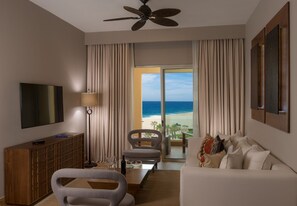 A hotel room with a large window offering a view of the beach, a ceiling fan, a flat-screen TV, a wooden cabinet, a sofa, and a coffee table.