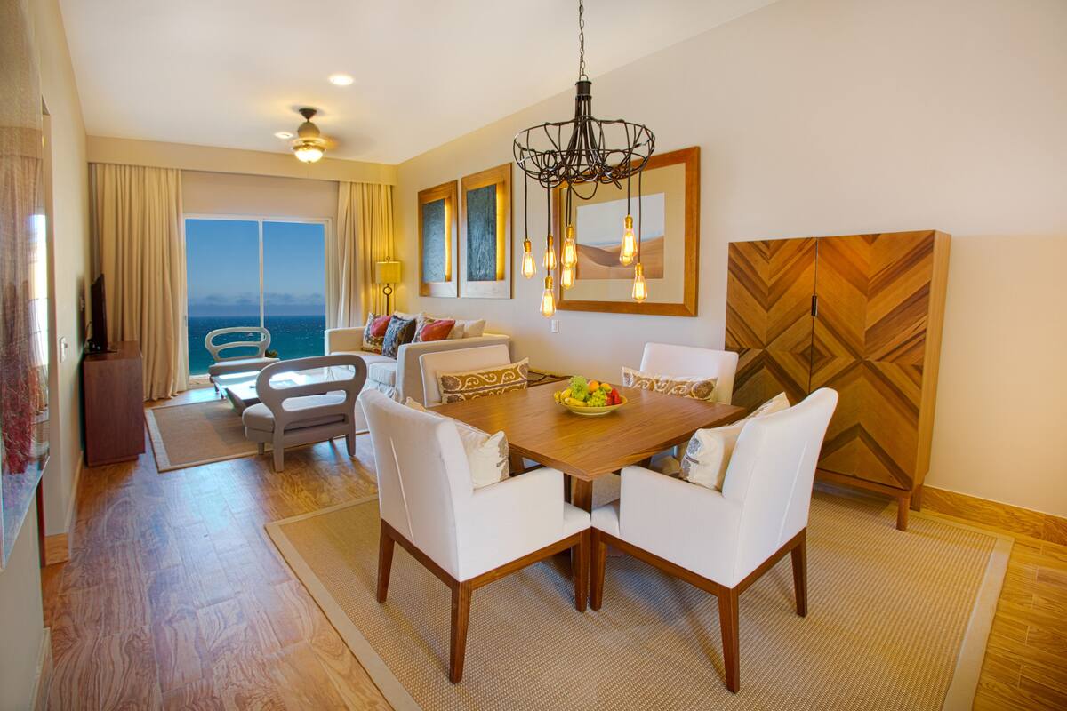A dining area with a wooden table, white chairs, and a chandelier. There is a sofa with cushions and a view of the sea through large windows.
