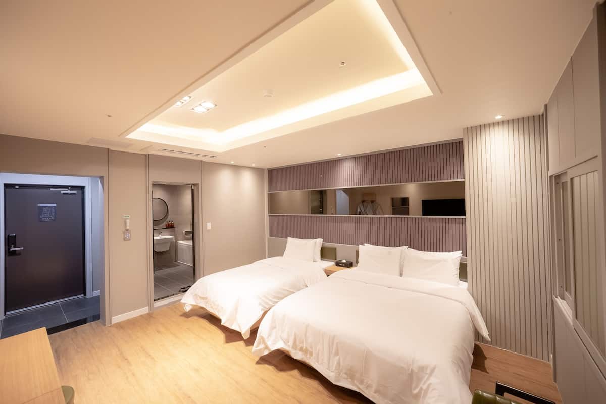 A modern hotel room with two beds, a built-in wardrobe, a vanity with a mirror, and a ceiling light fixture.