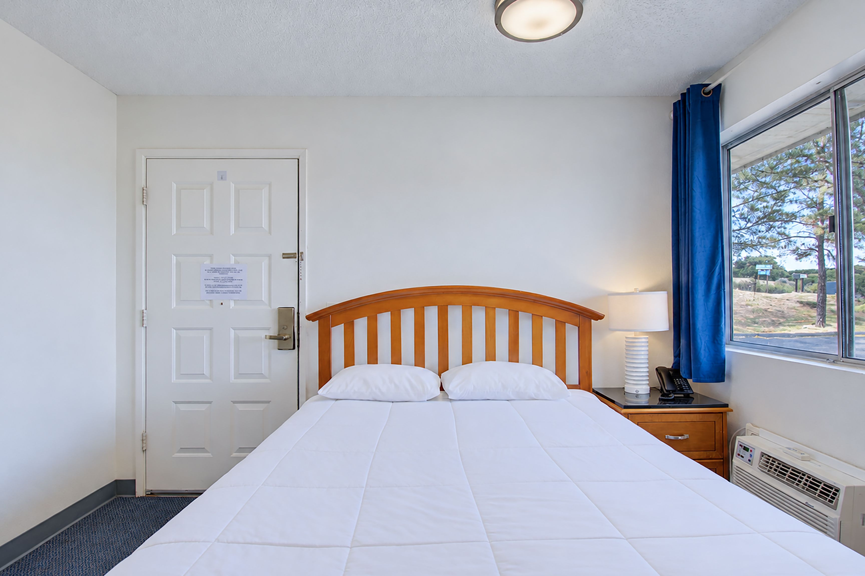 A hotel room with a bed, a door, a window with curtains, a bedside table with a lamp, and a thermostat.