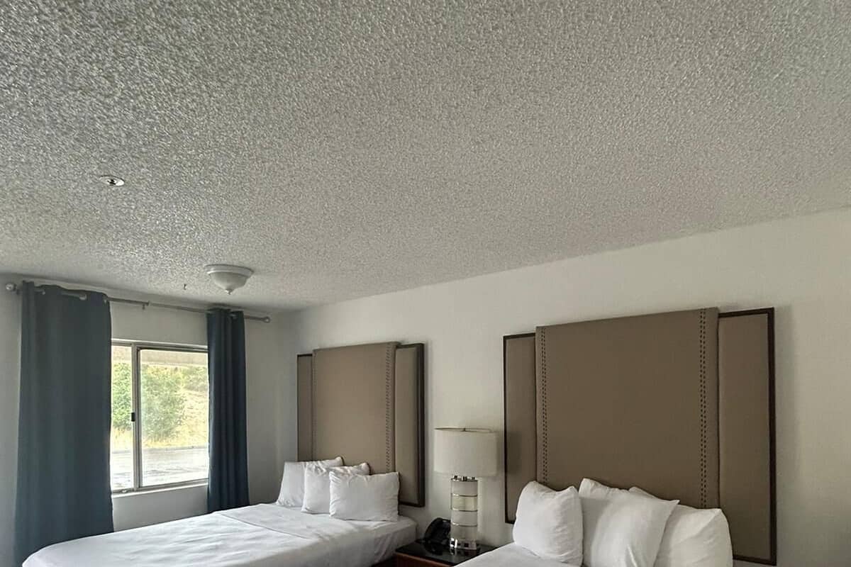 A hotel room with two beds, a bedside table with a lamp, and a window with curtains.