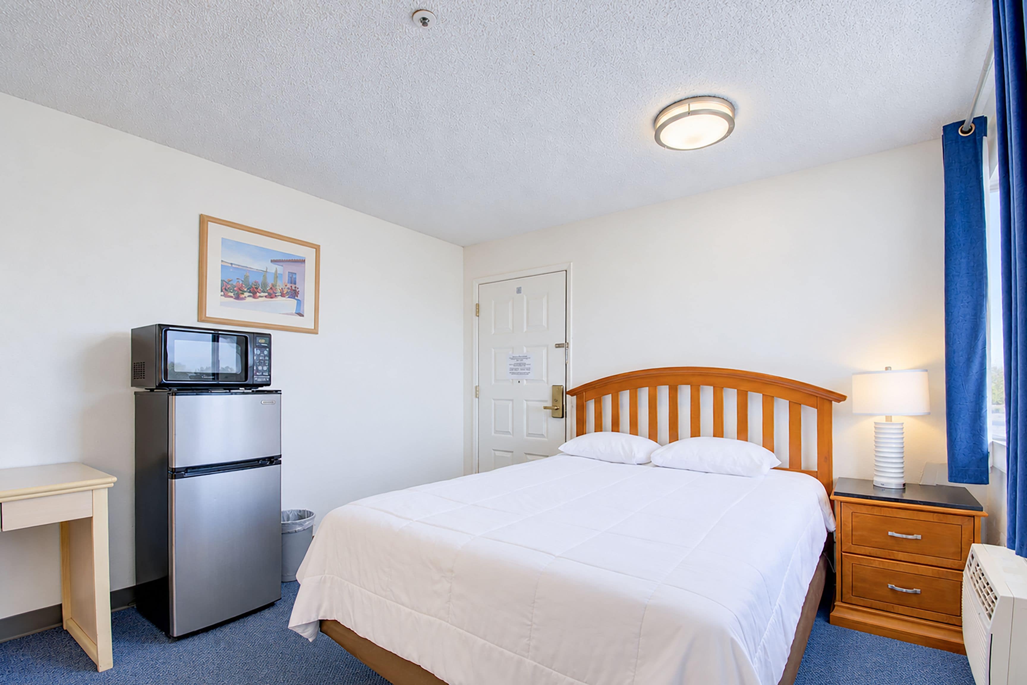Habitación de hotel con cama, escritorio con refrigerador, microondas, lámpara y un cuadro en la pared.