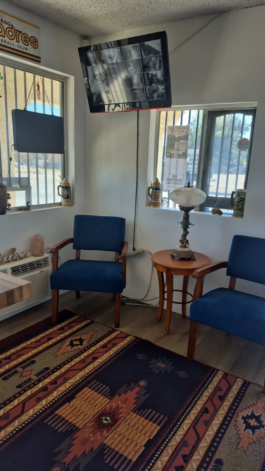Una habitación con dos sillas azules, una mesita redonda con una lámpara, una alfombra estampada, un televisor montado en la pared y una ventana con vistas al exterior.