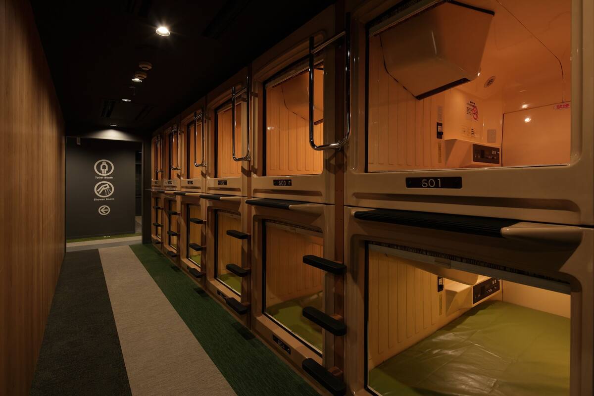 A row of wooden en-suite rooms with individual numbers, a green carpeted floor, and a wall-mounted sign indicating amenities.