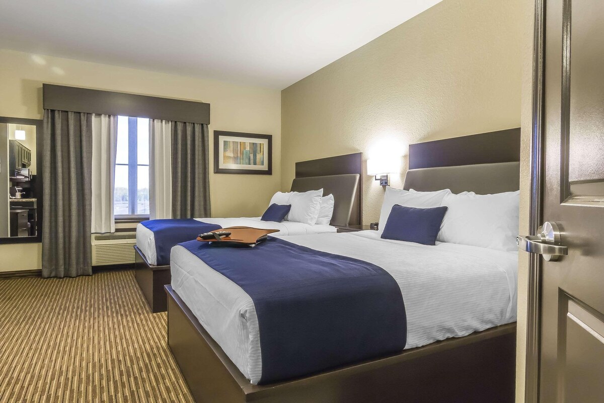 A hotel room with two beds, a desk, a chair, a window with curtains, a framed picture, and a wall-mounted lamp.
