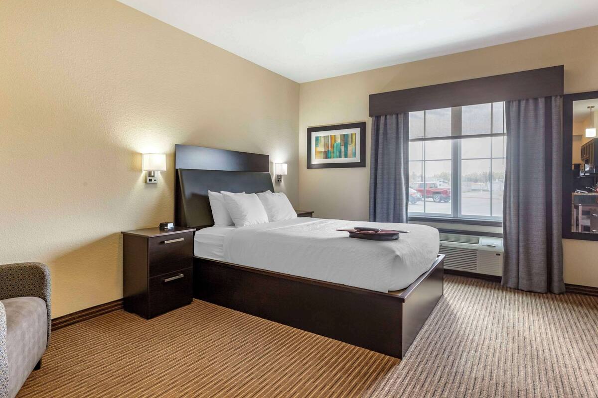 Une chambre d’hôtel avec un grand lit, une table de chevet, une lampe, un tableau encadré et une fenêtre avec des rideaux.