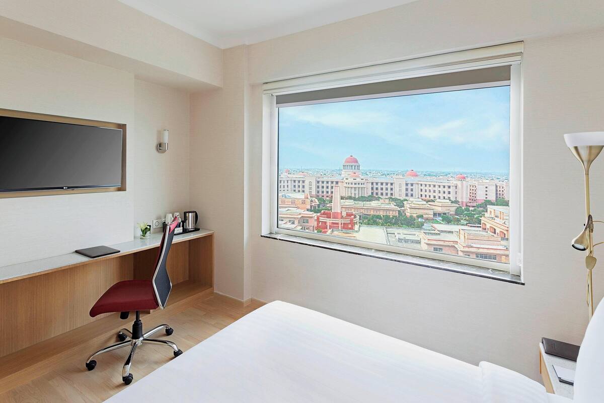 A hotel room with a large window, a desk with a chair, a bed, and a TV mounted on the wall.