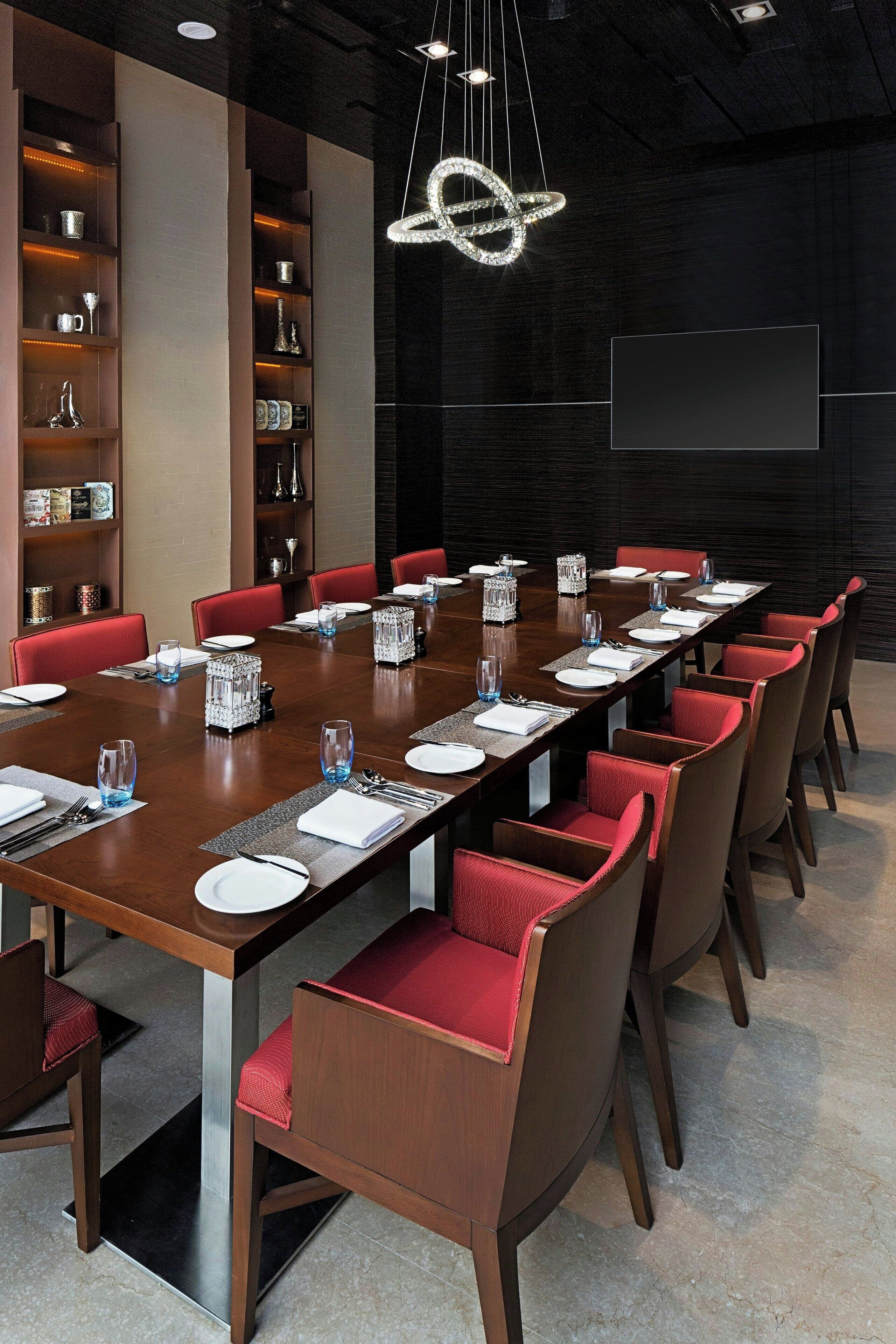Un salón de conferencias con una larga mesa preparada para una reunión, rodeado de sillas rojas, y un candelabro colgando en el techo.