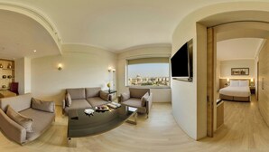 A modern hotel room with a curved sofa set, a coffee table, a flat-screen TV mounted on the wall, and a large window offering a city view. - Fairfield By Marriott Lucknow (Lucknow)