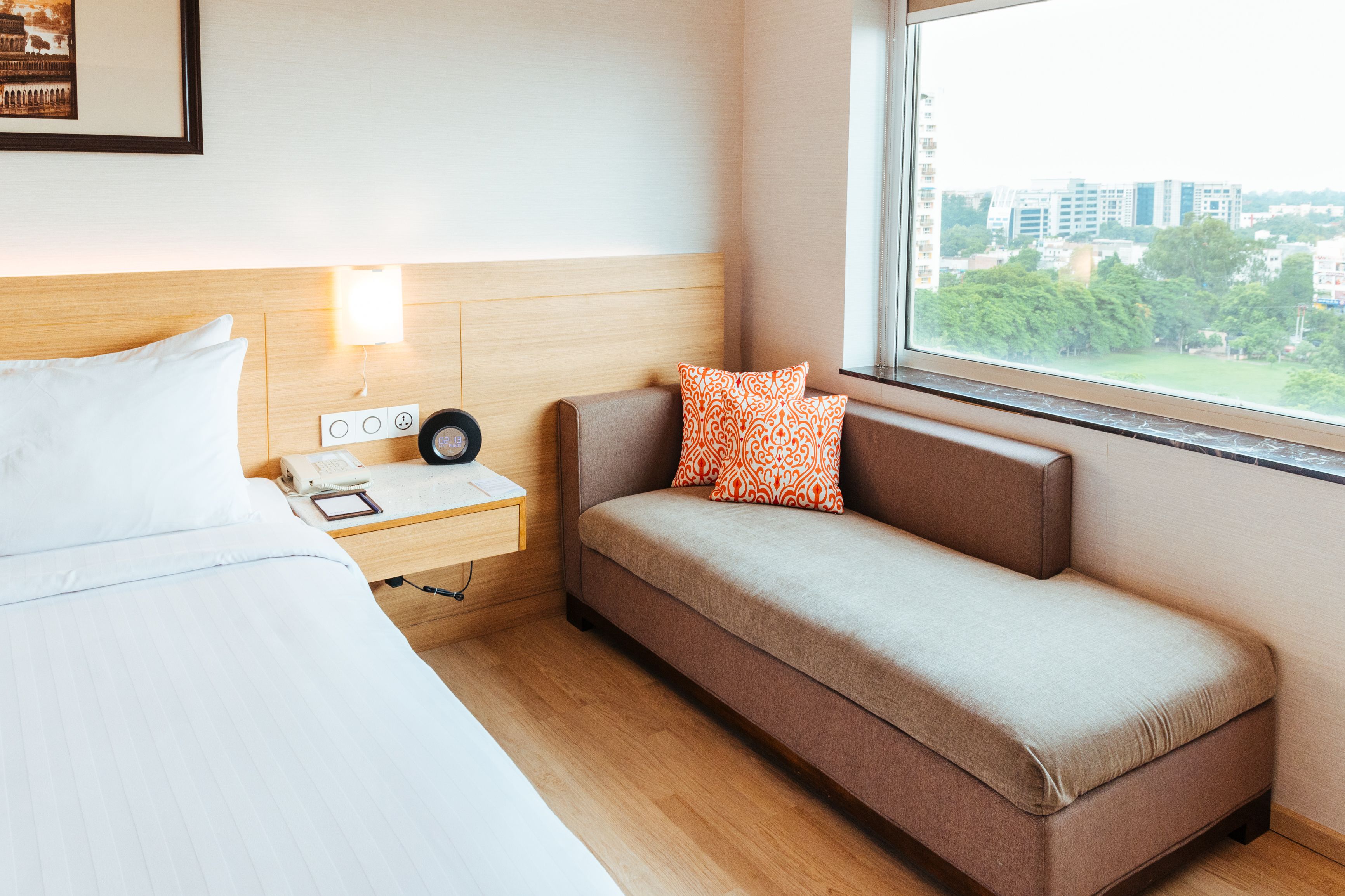 Une chambre d’hôtel avec un lit, une table de chevet avec un téléphone et une lampe, un canapé avec des coussins à motifs et une fenêtre donnant sur la ville.