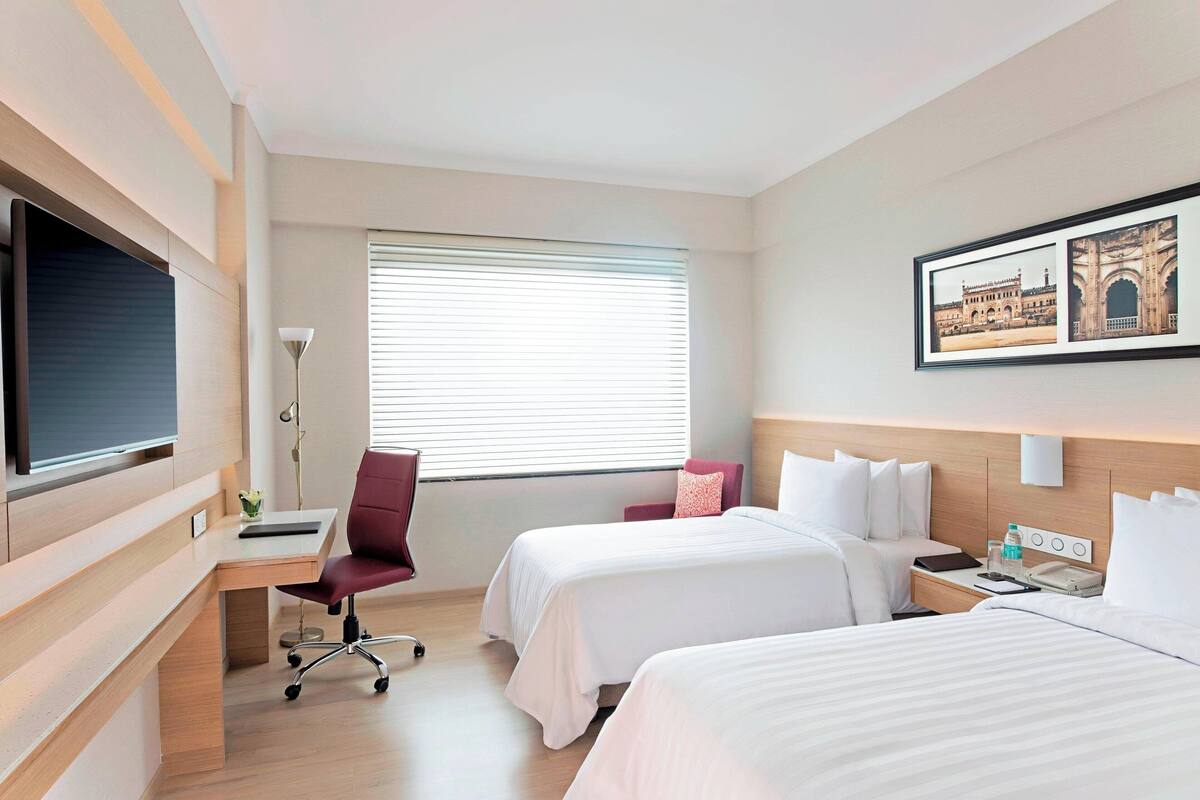 A hotel room with two beds, a desk, a chair, a TV, a window with blinds, and a wall-mounted light.