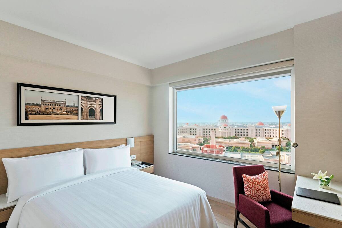 A hotel room with a large bed, a desk, a chair, a nightstand, and a window with a city view.