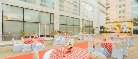 Un espacio para eventos al aire libre con mesas redondas, sillas blancas y un escenario con decoración floral.