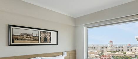 A hotel room with a large window, a bed with white linens, a framed picture of a building, and a nightstand with a phone and a book.