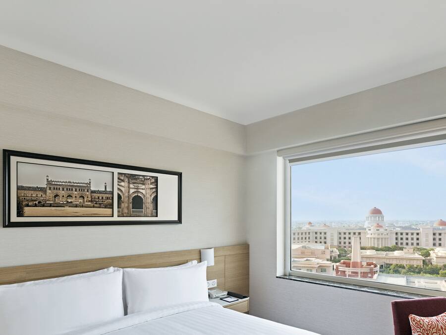 A hotel room with a large window, a bed with white linens, a framed picture of a building, and a nightstand with a phone and a book.