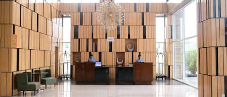A modern reception area with a high ceiling, wooden panel walls, and a central chandelier. There are two reception desks with computers, and green upholstered chairs to the left.