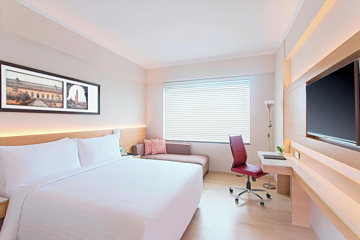 A hotel room with a large bed, a desk, a chair, a TV, and a window with blinds.