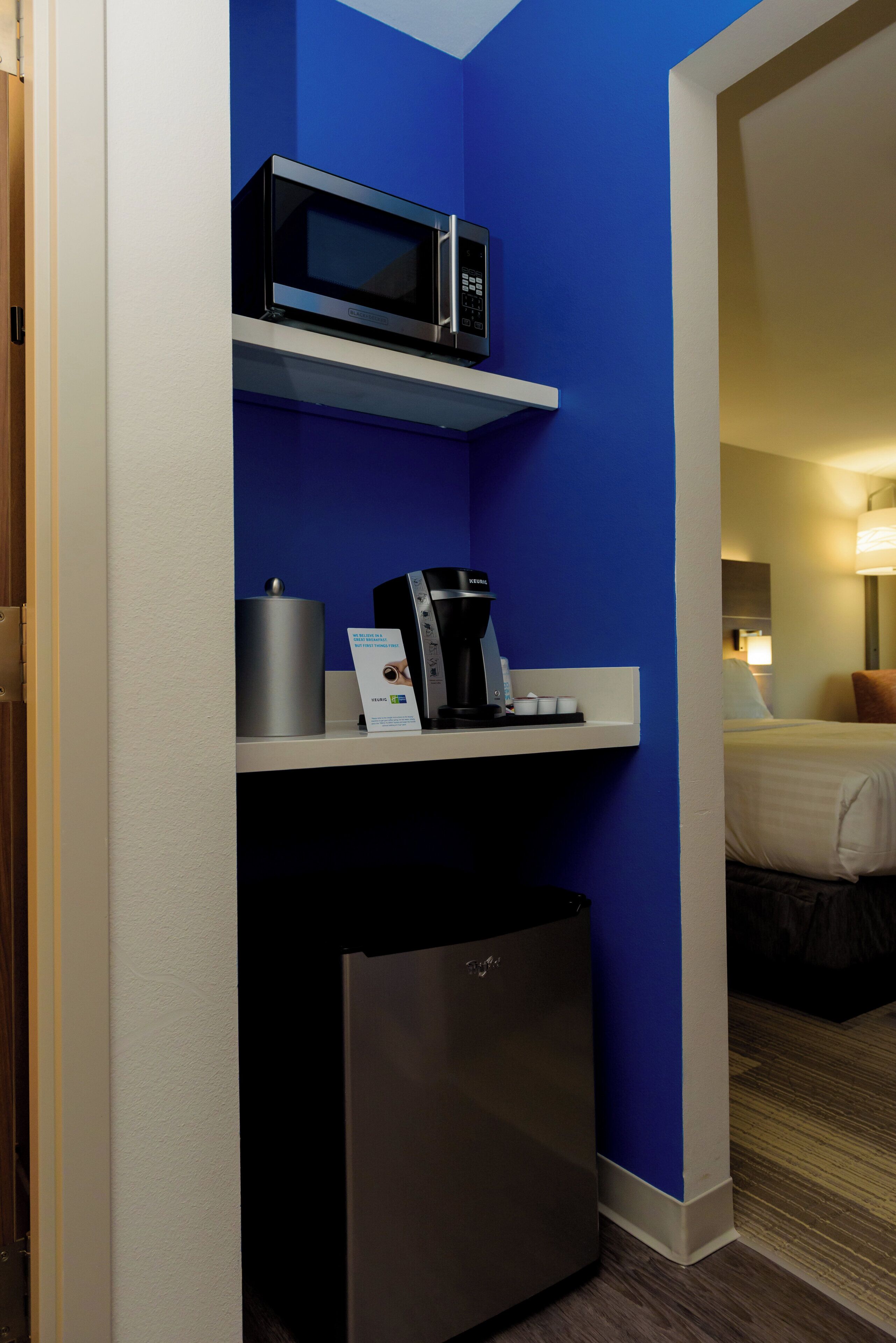 Une chambre d’hôtel compacte, dotée d’un four à micro-ondes, d’une machine à café et d’un mini-réfrigérateur, le tout placé sur une étagère blanche adossée à un mur bleu.