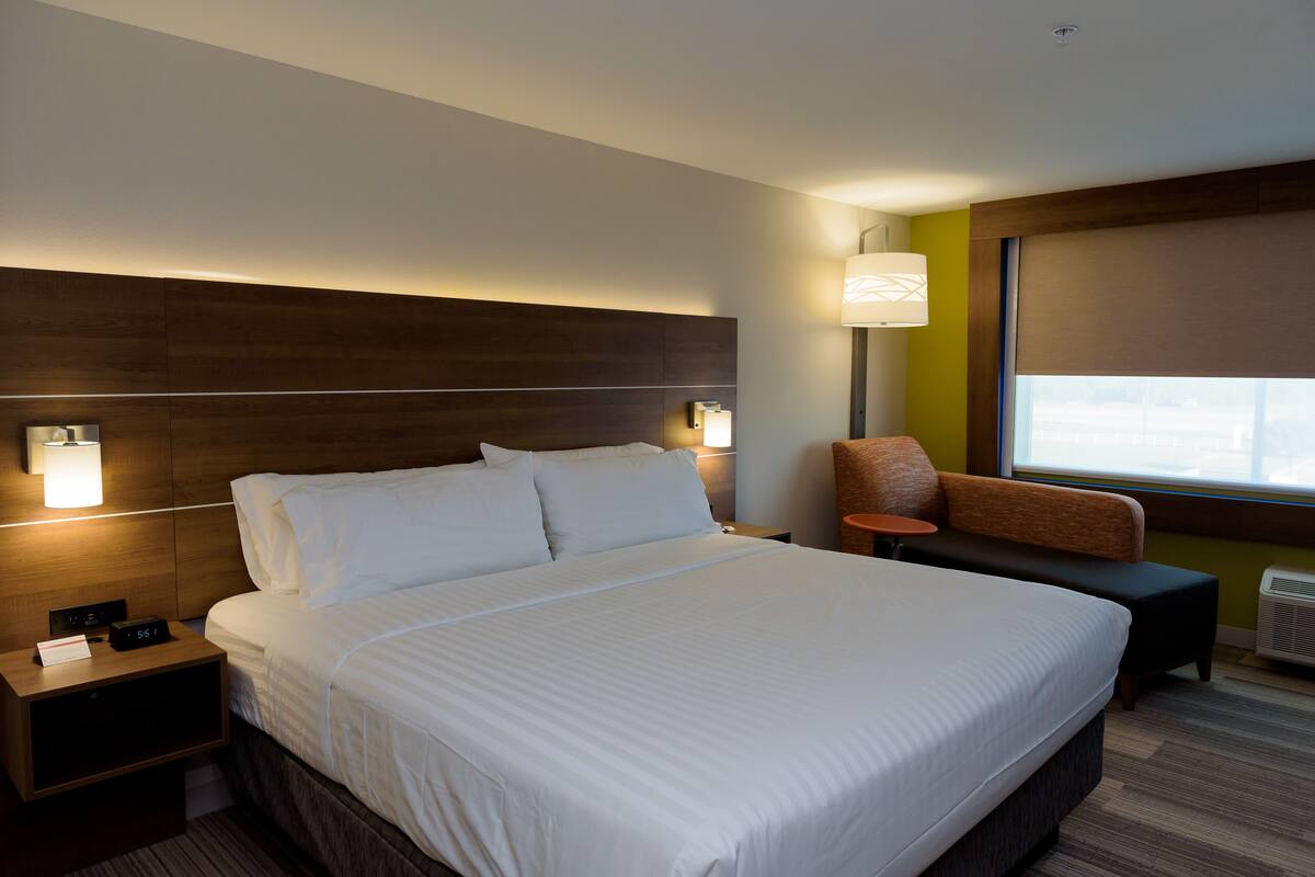 A hotel room with a large bed, a nightstand with a clock, a lamp, a sofa, and a window with a view.