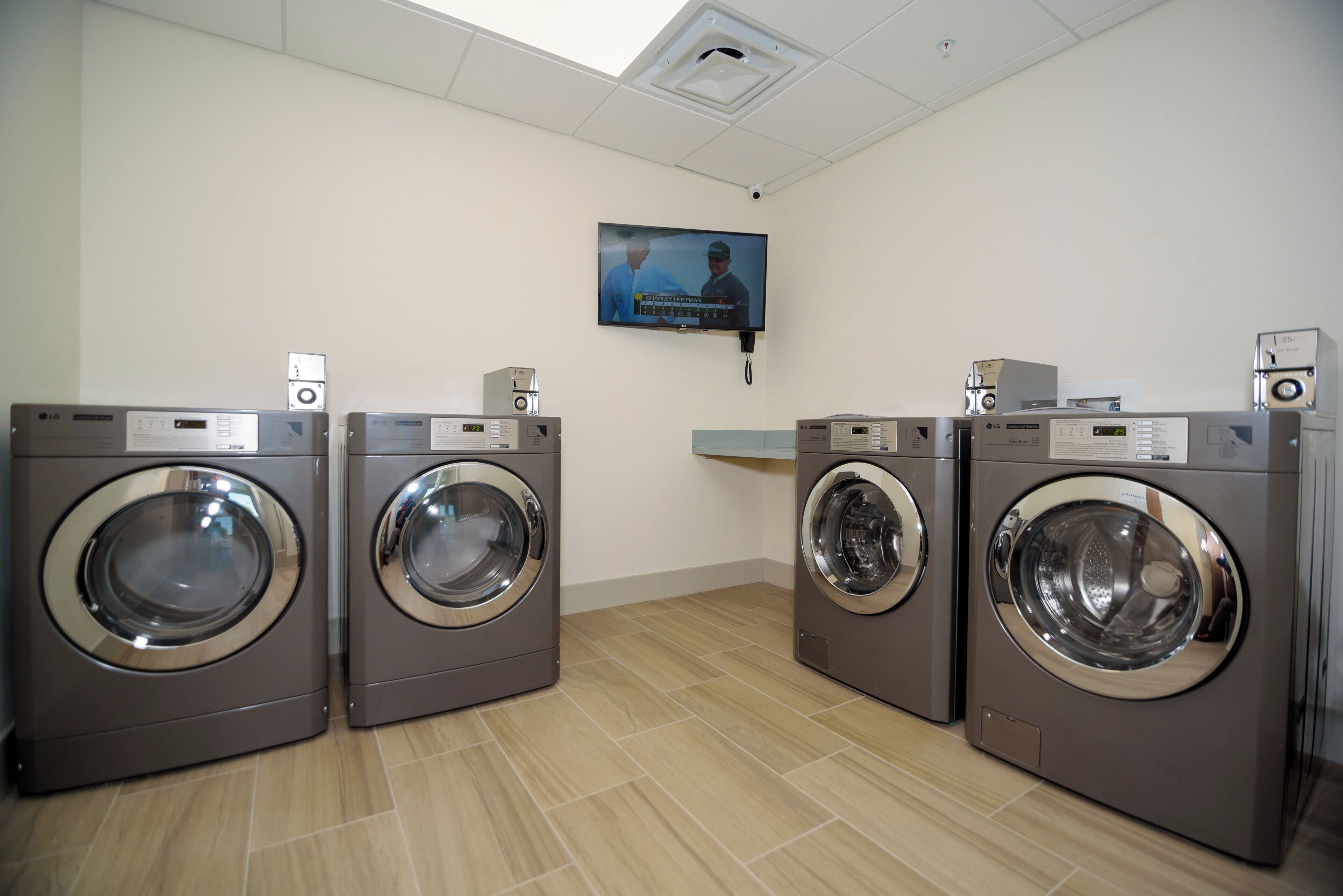 A laundry room with four front-loading washing machines, a flat-screen TV mounted on the wall, and a ceiling vent.