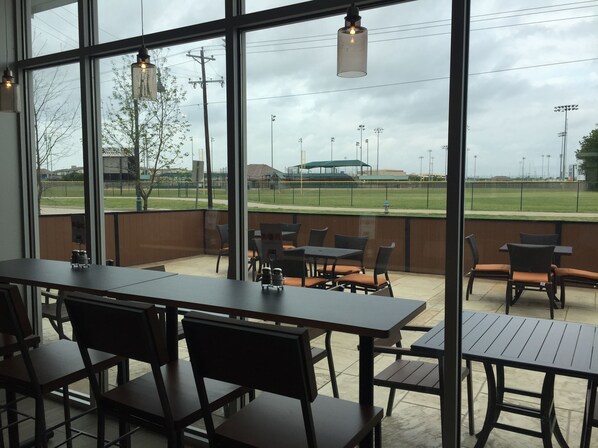 An indoor seating area with tables and chairs, large windows offering views of a sports field and stadium. - Holiday Inn Express & Suites McKinney - Frisco East by IHG (McKinney)