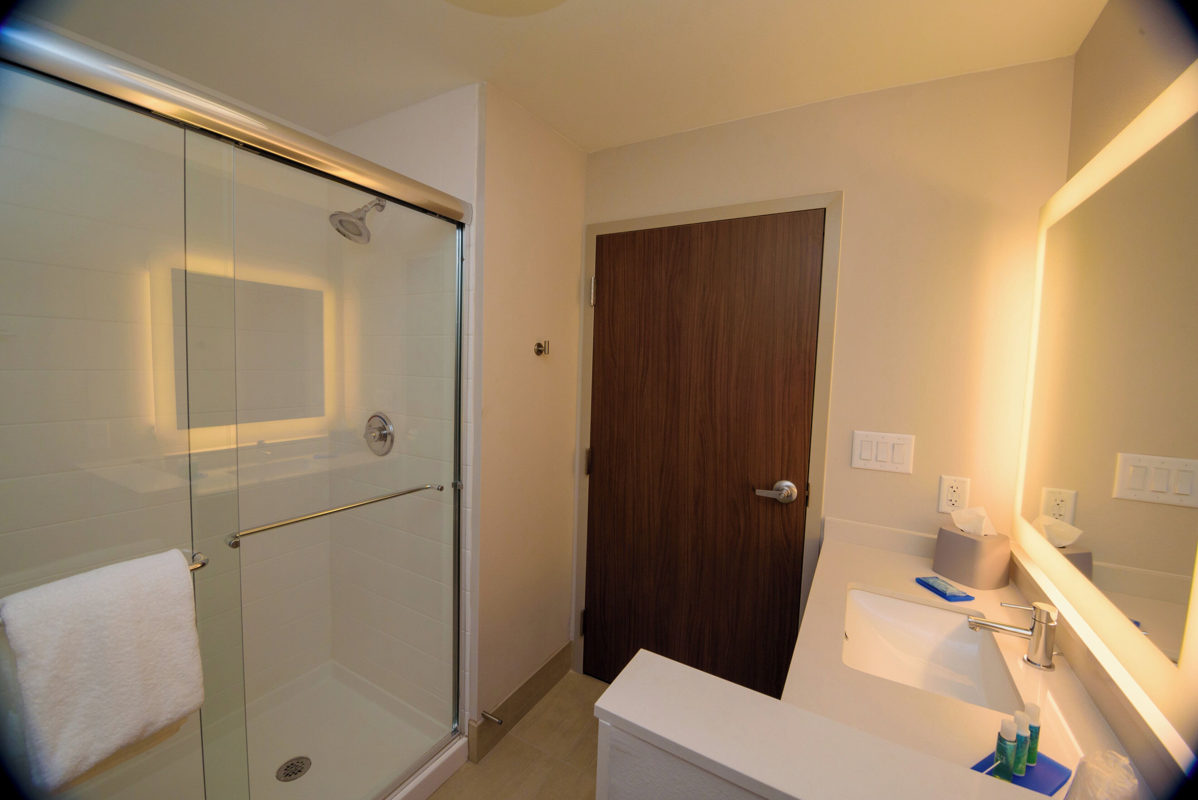A bathroom with a glass shower enclosure, a wooden door, a white sink with a mirror, and a towel on the floor.