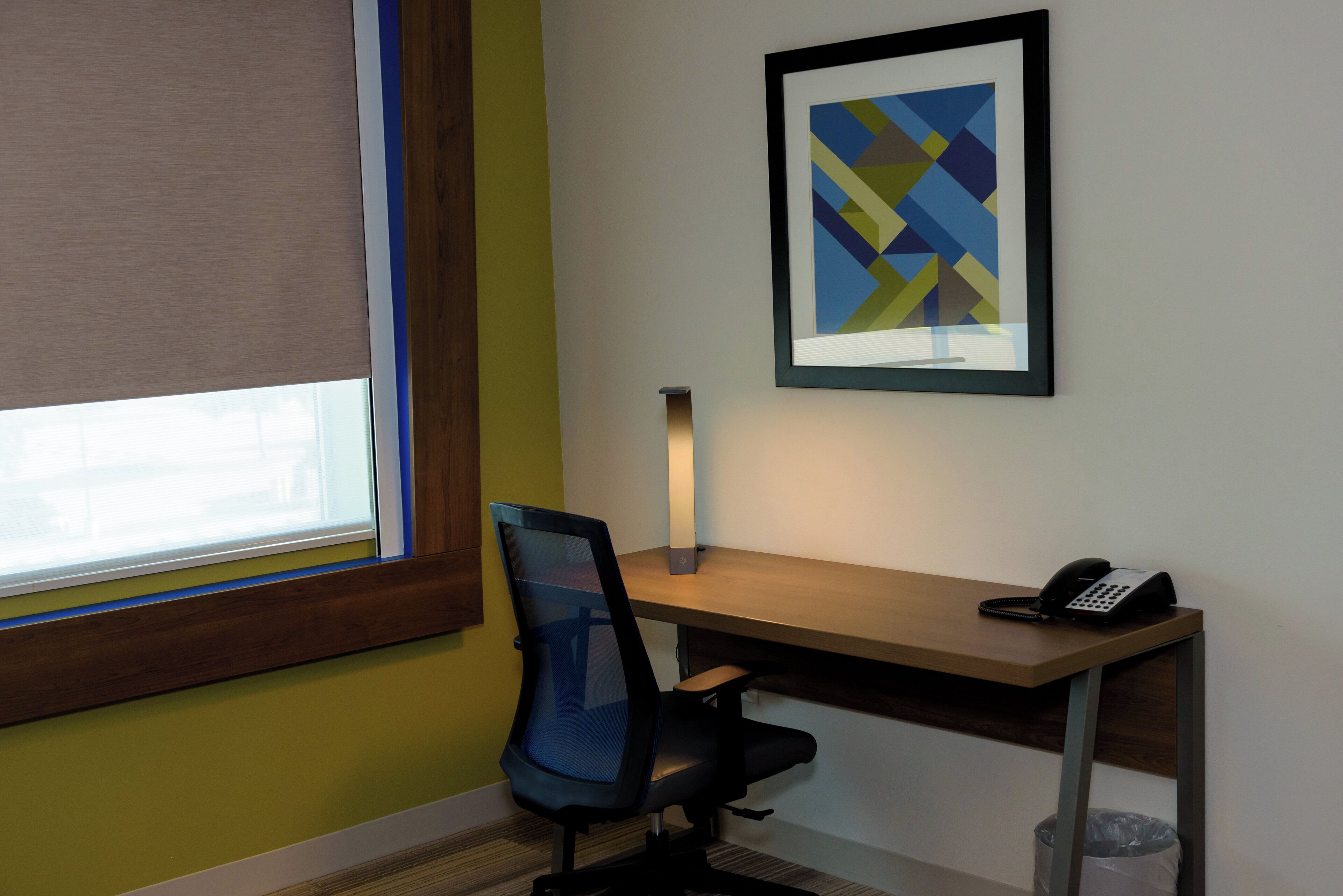 Une pièce comprenant un bureau, une chaise, une fenêtre avec des stores, un tableau abstrait encadré et un téléphone.