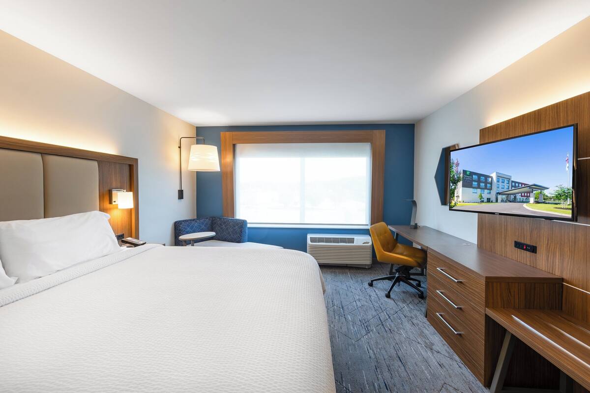 A hotel room with a large bed, a desk, a chair, a TV, and a window with a view of buildings.