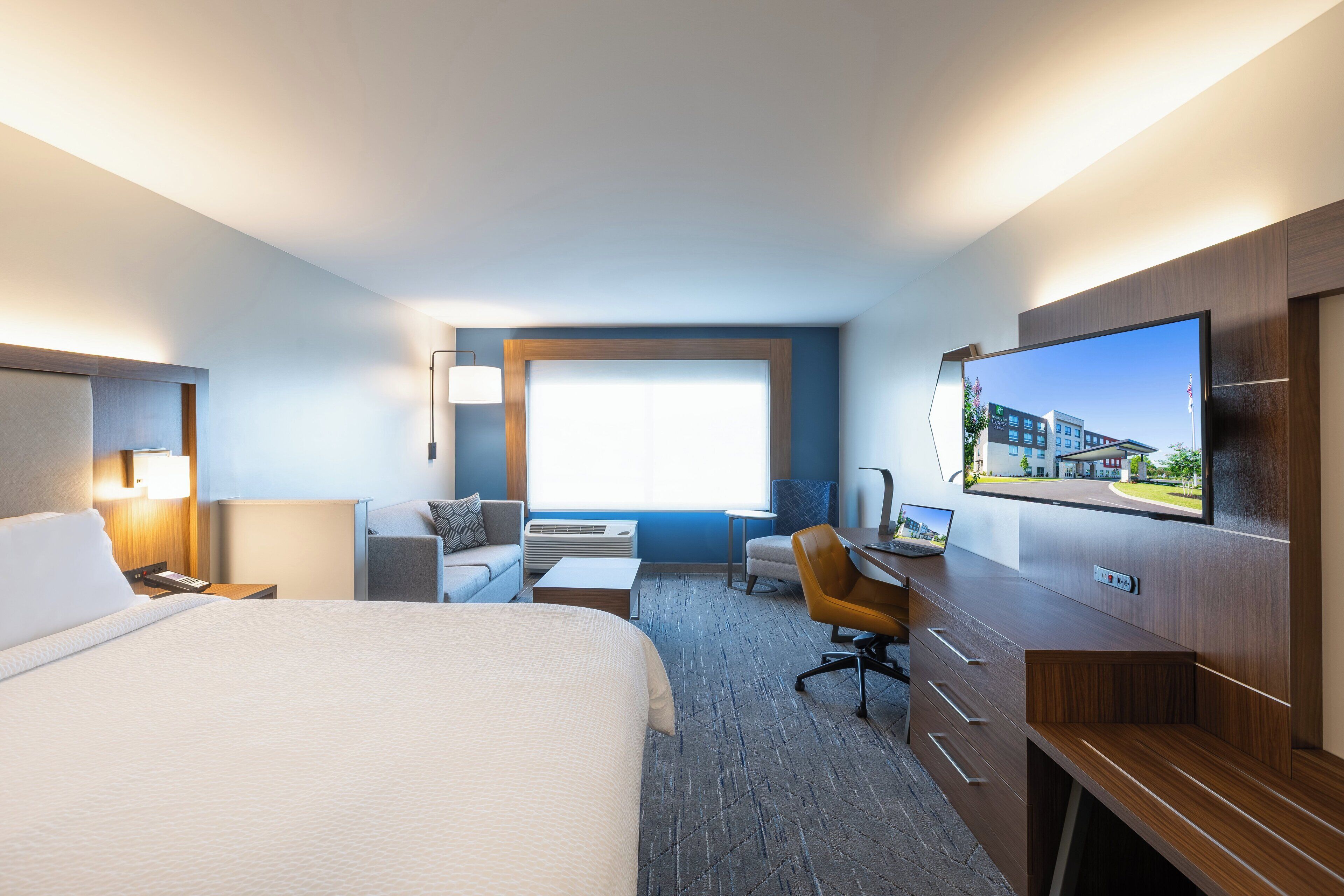 A hotel room with a large bed, a desk, a chair, a TV, and a window with a view of buildings.