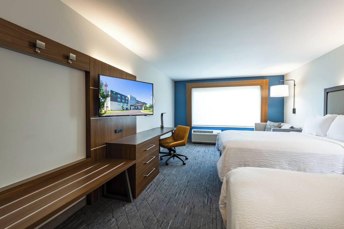 A hotel room with a large bed, a desk with a chair, a television mounted on the wall, and a window providing a view of the outside.