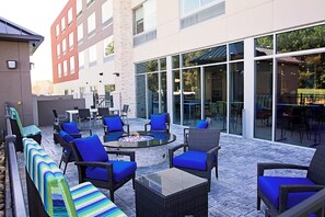 An outdoor seating area with wicker furniture and blue cushions, a fire pit, and a glass wall overlooking a building. - Holiday Inn Express & Suites Greenwood Mall by IHG (Greenwood)