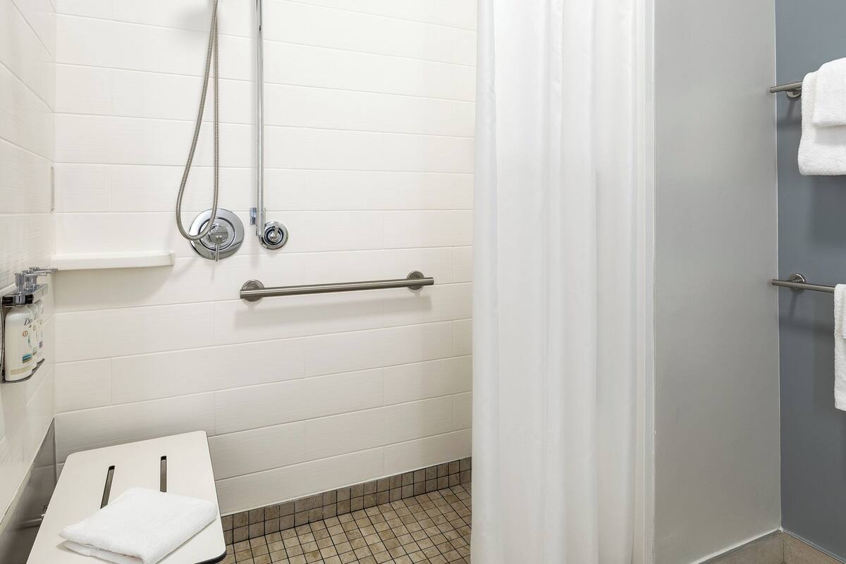 A bathroom with white tiles, a shower with a handheld showerhead, a grab bar, and two towel racks with towels.