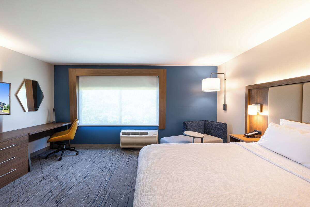 A hotel room with a bed, a desk, a chair, a small table, a lamp, and a window.