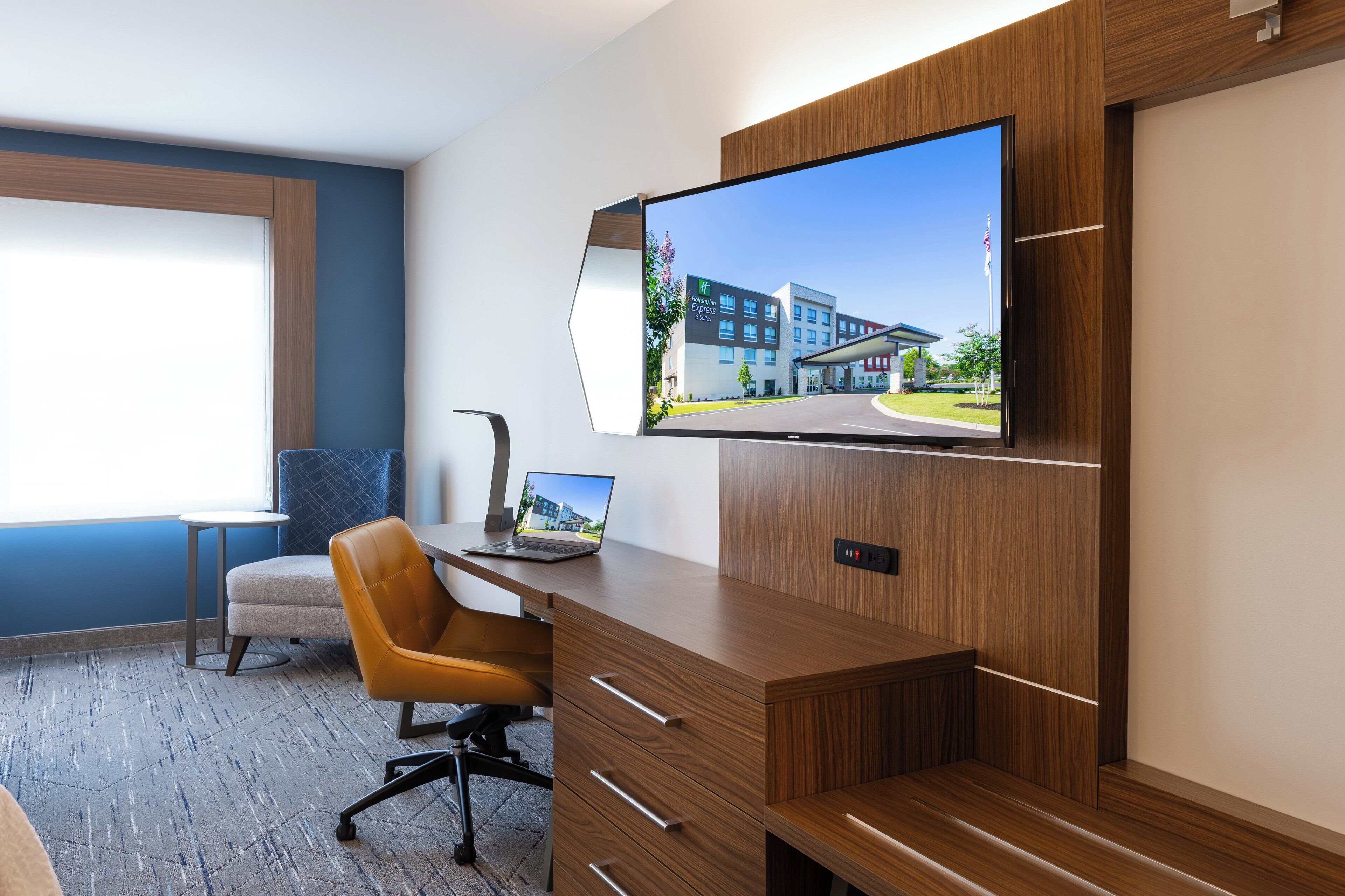 A modern hotel room with a wooden desk, a laptop, a flat-screen TV mounted on the wall, and a comfortable chair.