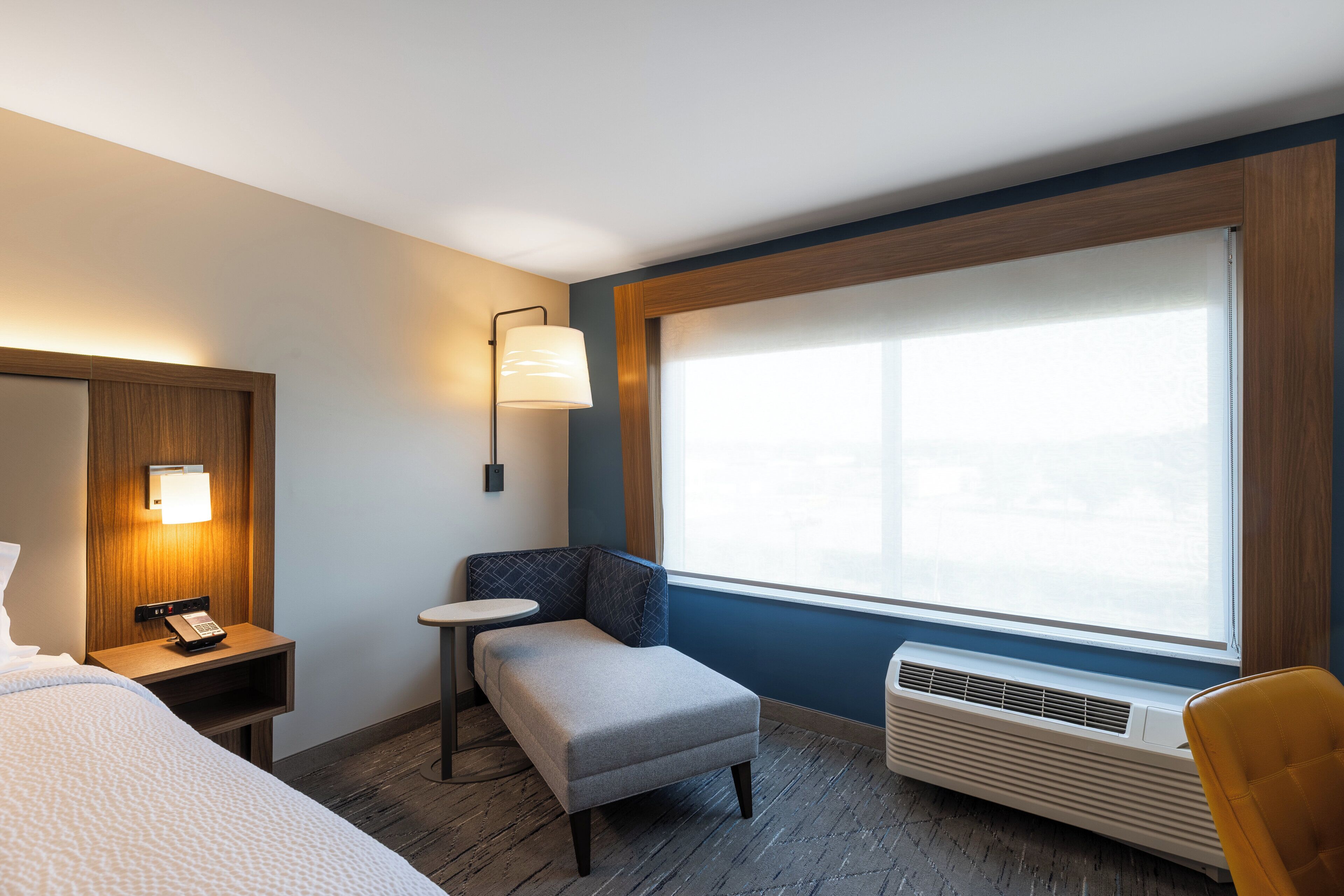 Habitación de hotel con cama, una mesita, una silla, escritorio, lámpara, calefactor y ventana grande.