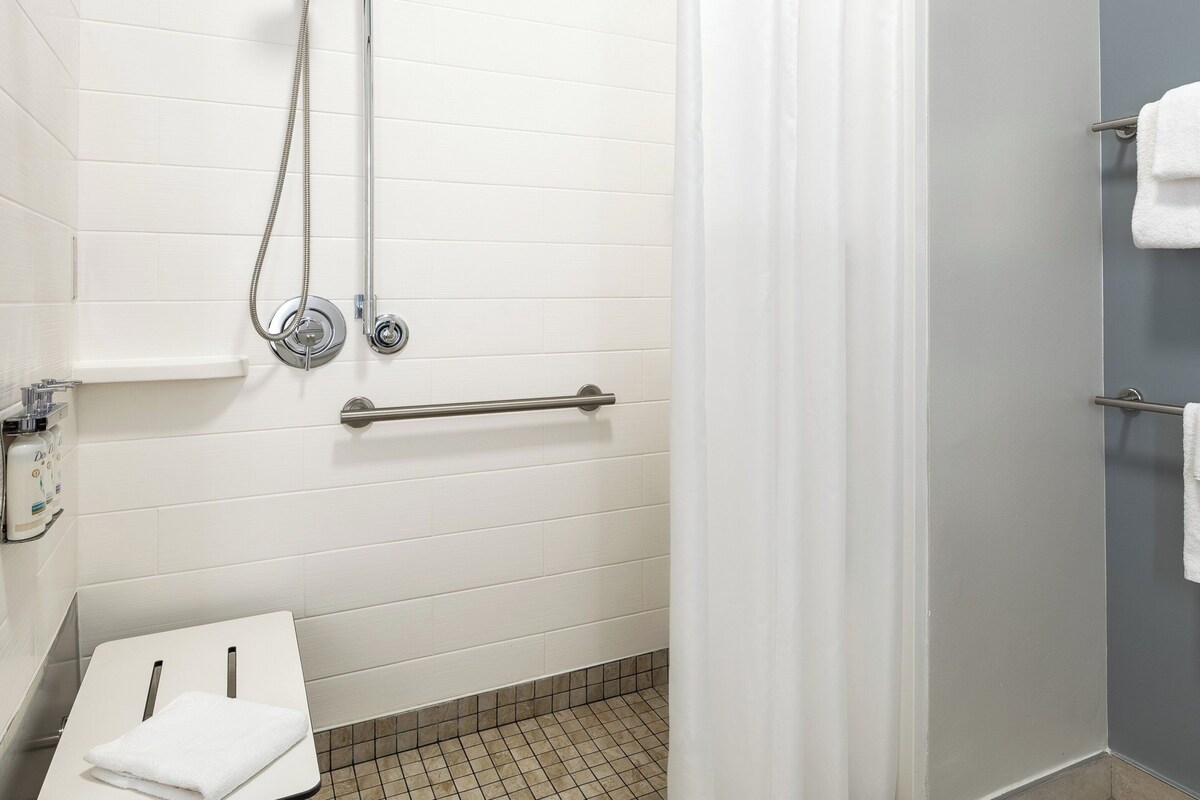Un baño con baldosas blancas, una ducha con cabezal de ducha manual, un asidero y dos barras para toallas con toallas.