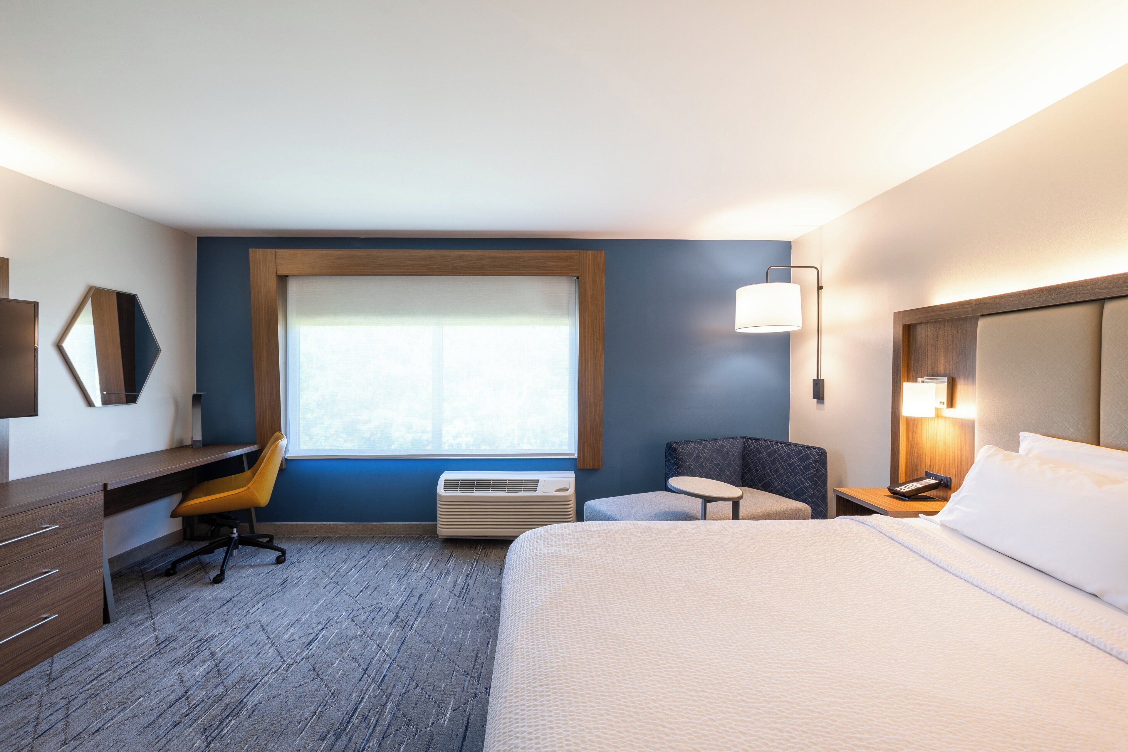 A hotel room with a bed, a desk, a chair, a window, and a wall-mounted lamp.