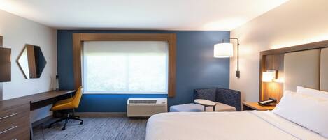 A hotel room with a bed, a desk, a chair, a window, and a wall-mounted lamp.