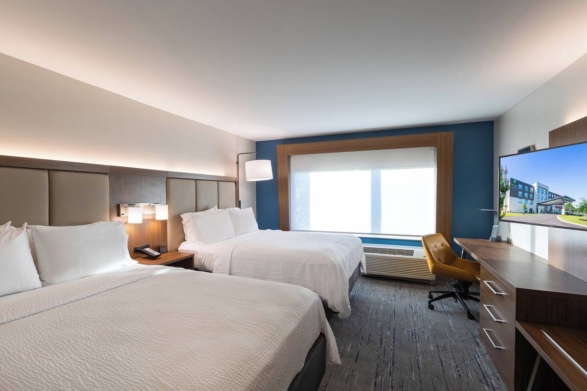 A hotel room with two beds, a desk, a chair, a TV, and a large window.