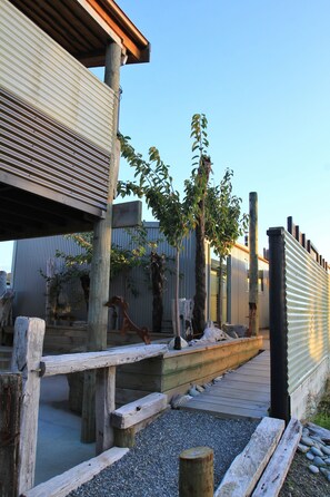 A wooden deck with a metal railing, surrounded by gravel and small plants. - Code Time Lodge (Okarito)