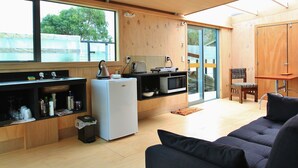 Een compacte keuken met een koelkast, magnetron en gootsteen. Er is een zithoek met een stoel en een bank. Grote ramen bieden natuurlijk licht en uitzicht op de omgeving.