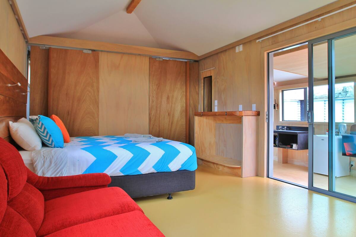 A modern bedroom with a bed, a red sofa, and a sliding glass door leading to a kitchen area.