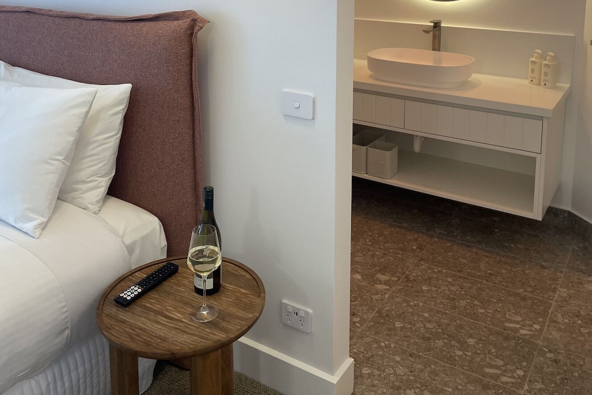 Habitación de hotel con cama, una mesa redonda con un control remoto y una botella de vino, una lámpara de pared y un baño con un espejo redondo y un lavabo blanco.