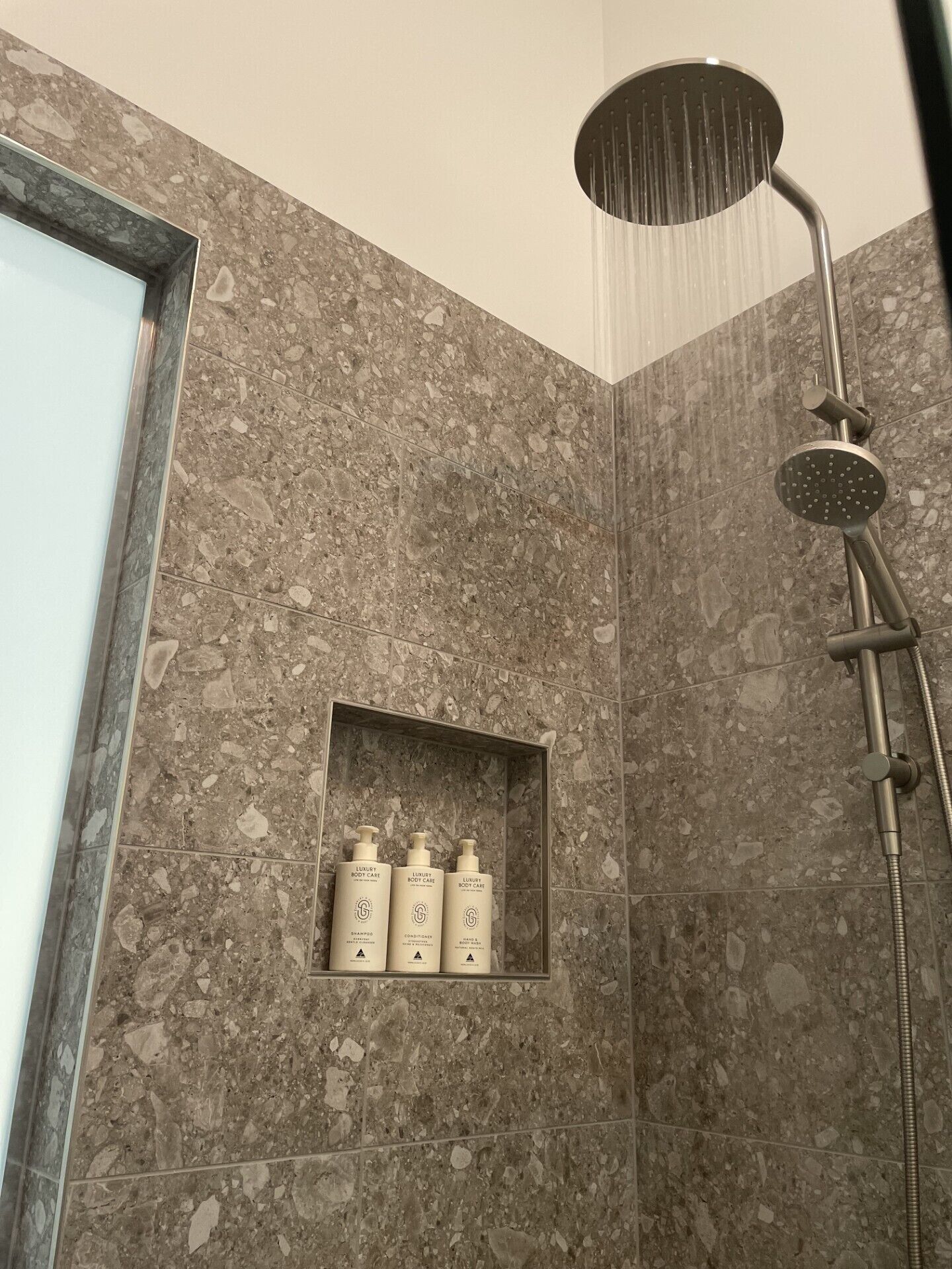 A shower with a rain head, a marble wall, and a shelf with three bottles.