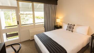 A hotel room with a large bed, a desk, a chair, a nightstand, a lamp, and a view of the outdoors through a sliding door.