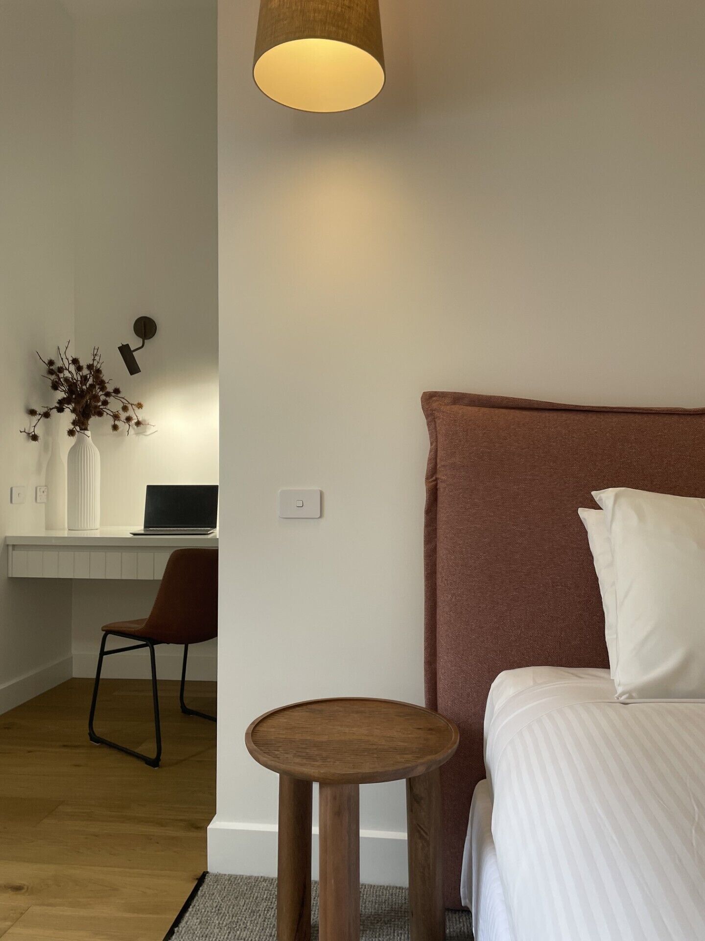 A bedroom with a bed, a desk with a laptop, a chair, a small round table, and a wall-mounted lamp.