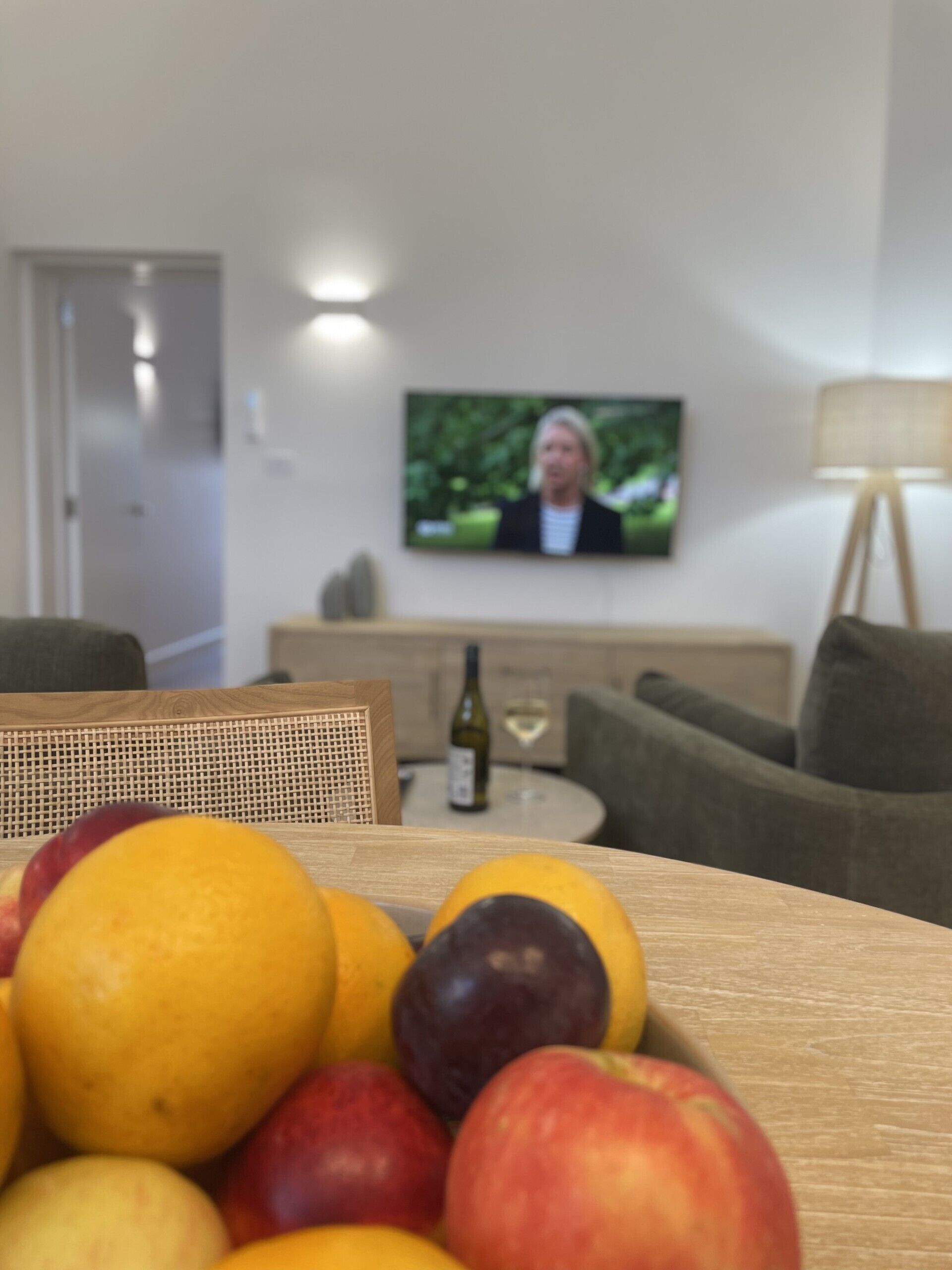 A dining table with a fruit bowl in the foreground and a television in the background.
