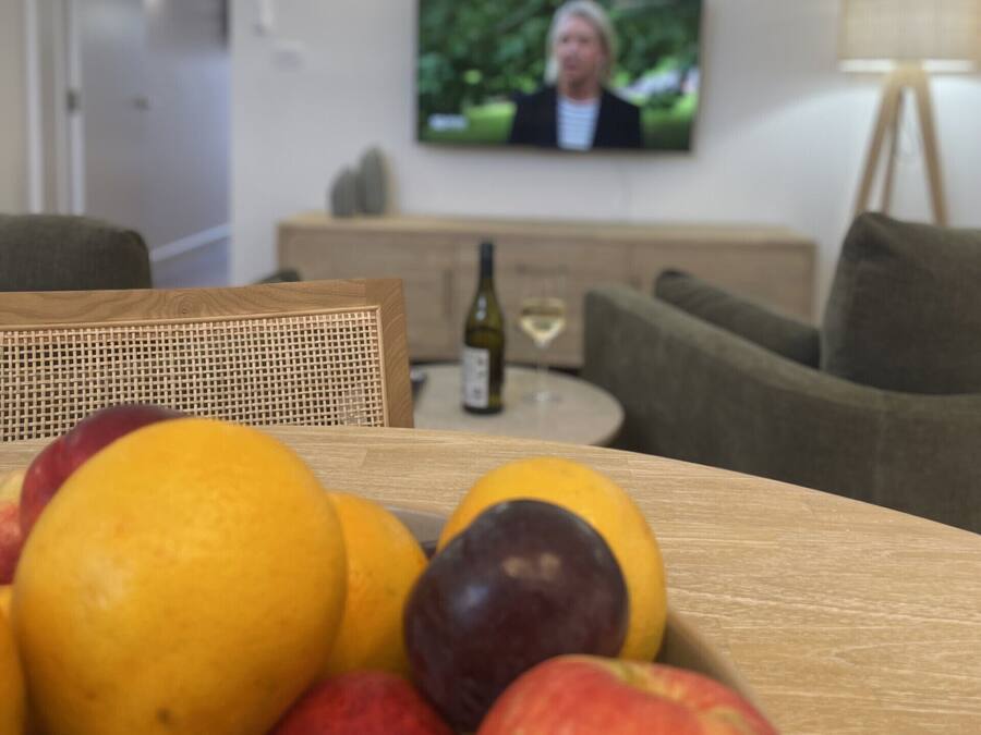 Une table à manger avec un panier de fruits au premier plan et un téléviseur à l’arrière-plan.