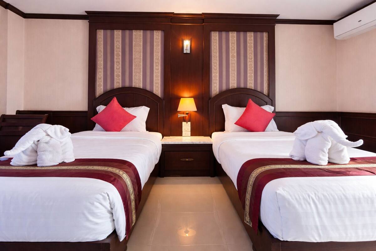 A hotel room with two beds, a nightstand with a lamp, and a wall-mounted air conditioner.